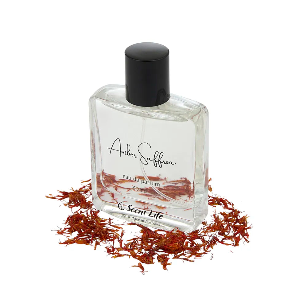 A clear Eau De Parfum bottle labeled "Amber Saffron" by Scent Life is positioned on a white background, accompanied by scattered saffron strands. It showcases a black cap, with the Scent Life branding prominently displayed at the bottom, suggesting delightful undertones of green tea jasmine within its sophisticated design.