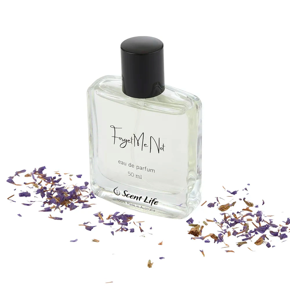 A clear glass bottle of Scent Life's Forget Me Not Eau de Parfum, 50 ml, with a black cap stands elegantly amidst scattered purple flower petals. The fragrance is enhanced by refreshing notes of bergamot.