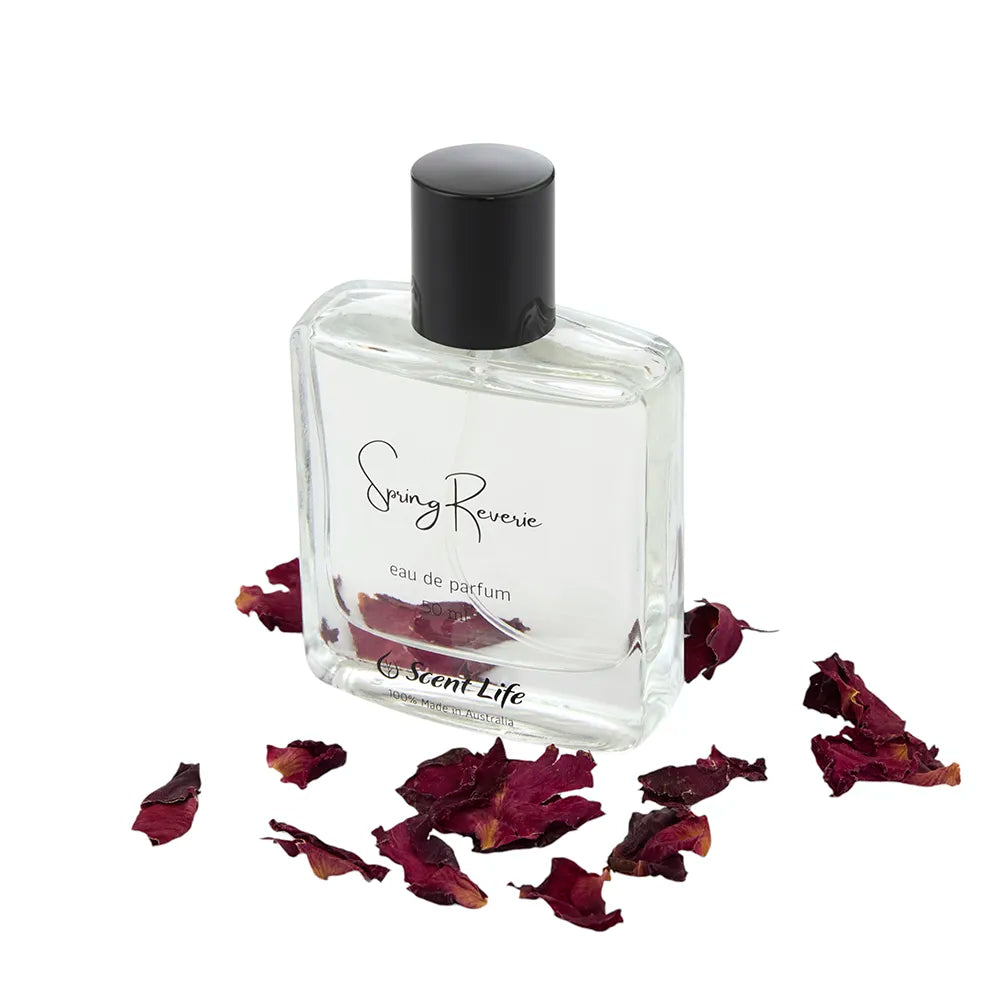 A clear glass perfume bottle from the Scent Life brand, labeled Spring Reverie eau de parfum and topped with a black cap, is elegantly surrounded by scattered dried red rose petals, capturing the essence of blooming cactus flowers.