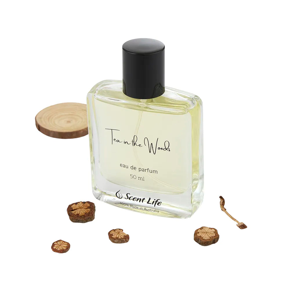 A 50 ml Eau de Parfum bottle of Scent Life's Tea in the Woods, renowned for its long-lasting fragrance, is displayed in a clear glass bottle with a sleek black cap. It elegantly sits next to small wooden slices against a pristine white background.