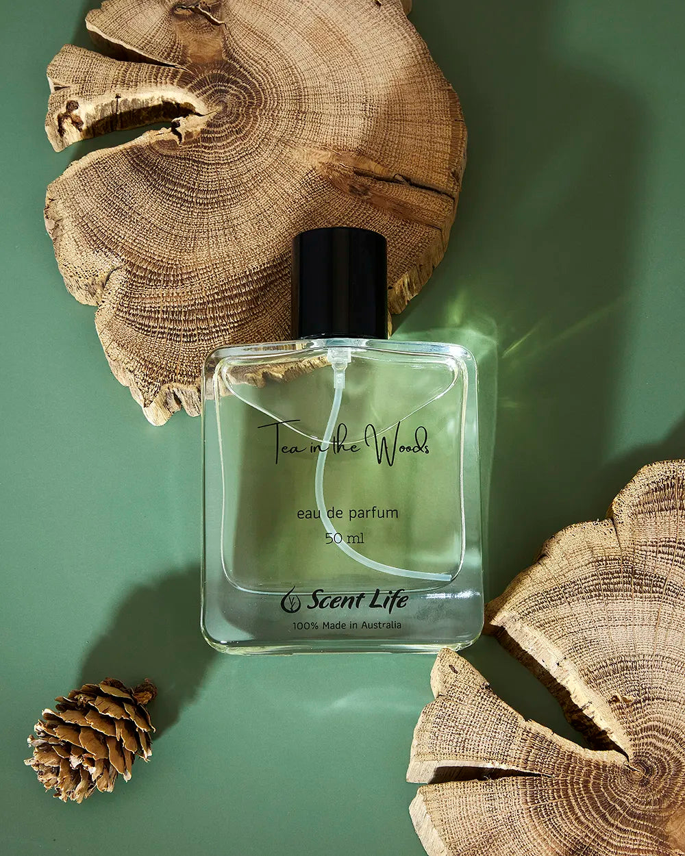 A bottle of Scent Life's Tea in the Woods eau de parfum sits atop a green surface, flanked by two sizable wooden slices and a small pine cone. This sophisticated rectangular bottle with its black cap offers a long-lasting scent that embodies the spirit of nature.