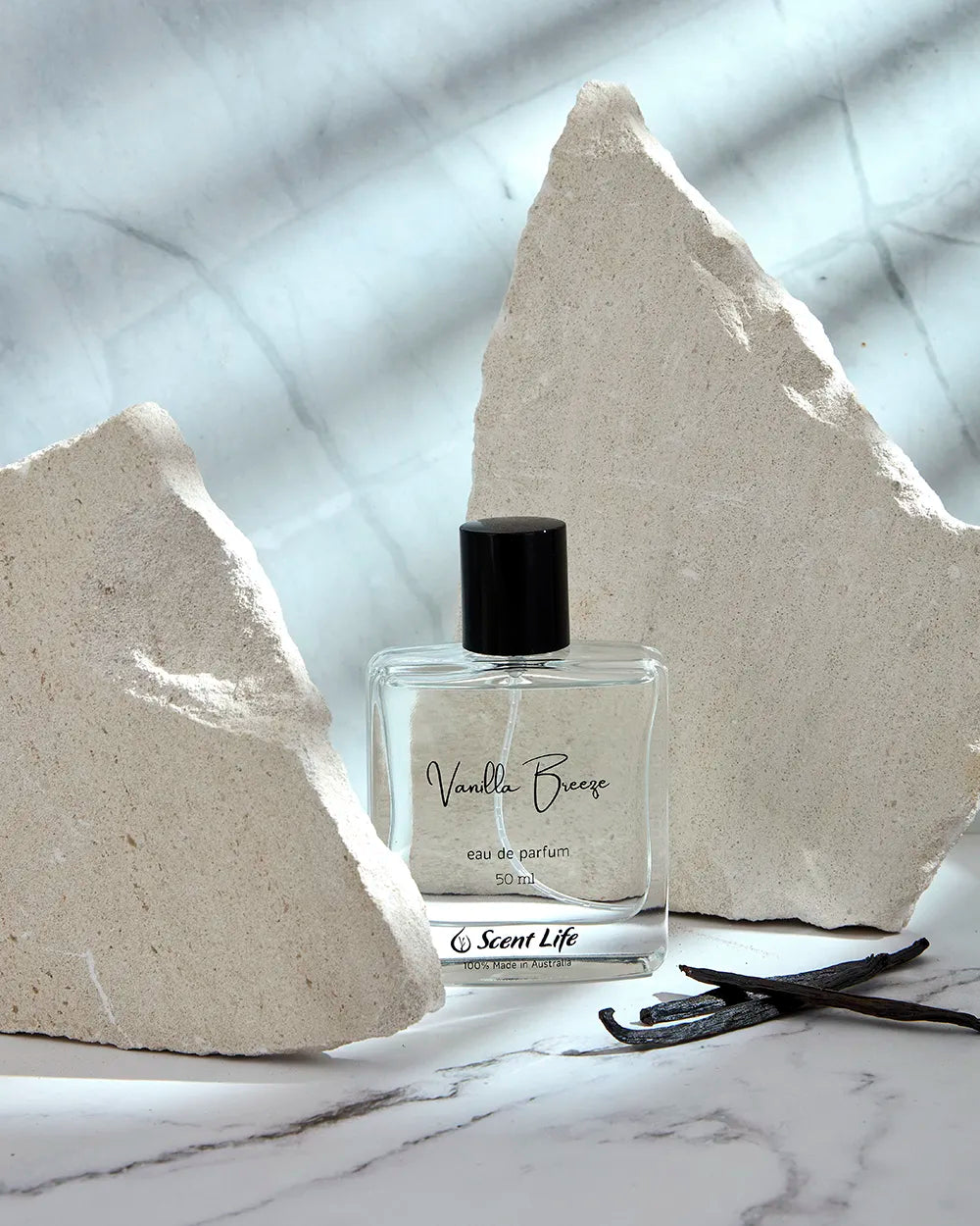 A bottle of Scent Life's Vanilla Breeze eau de parfum sits elegantly on a marble surface, encircled by large rock fragments. A vanilla bean rests close by, with natural light casting graceful shadows that amplify the fragrance's calm and sophisticated atmosphere.