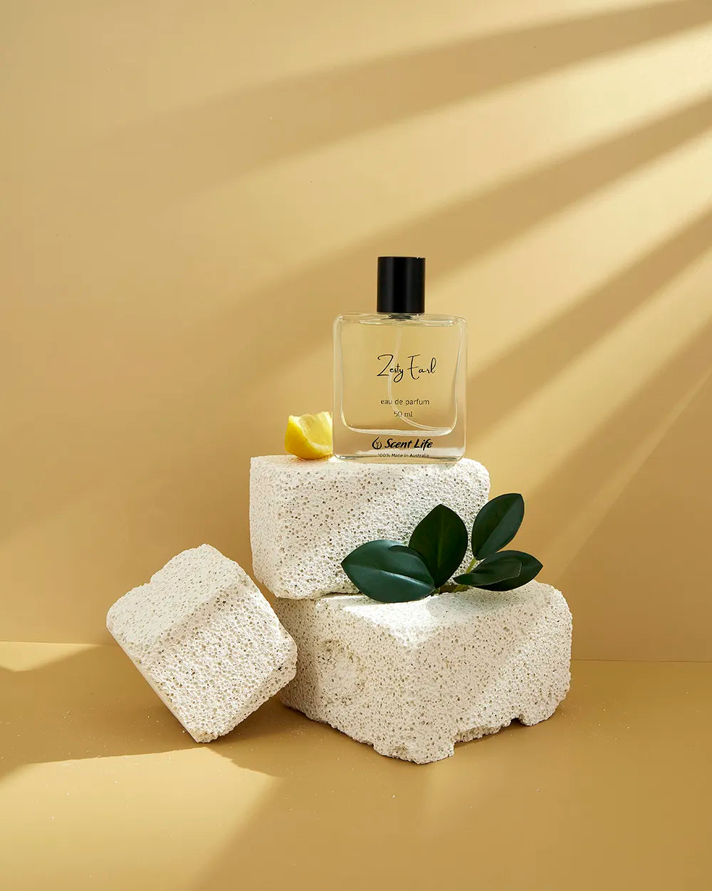 A bottle of Scent Life's Zesty Earl Eau de Parfum is elegantly perched on three textured white stones against a beige backdrop. Next to the bottle, a small yellow petal and green leaves enhance the scene, their soft shadows cast by warm light suggesting invigorating citrusy notes reminiscent of an Earl Grey tea experience.